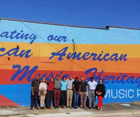 A diverse group of people stands in front of a colorful mural on the side of a brick building. The mural has blue, orange, and yellow stripes with the text "Celebrating our African American Music Heritage" on top.