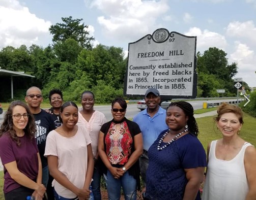 Group of smiling people of different races standing next to a road in front of a sign that says "Freedom Hill: Community established here by freed blacks in 1865. Incorporated as Princeville in 1885."