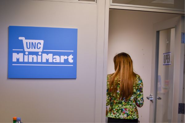 Sign that says UNC Mini Mart outside of a doorway where you can see the back of a woman who is entering the mini mart