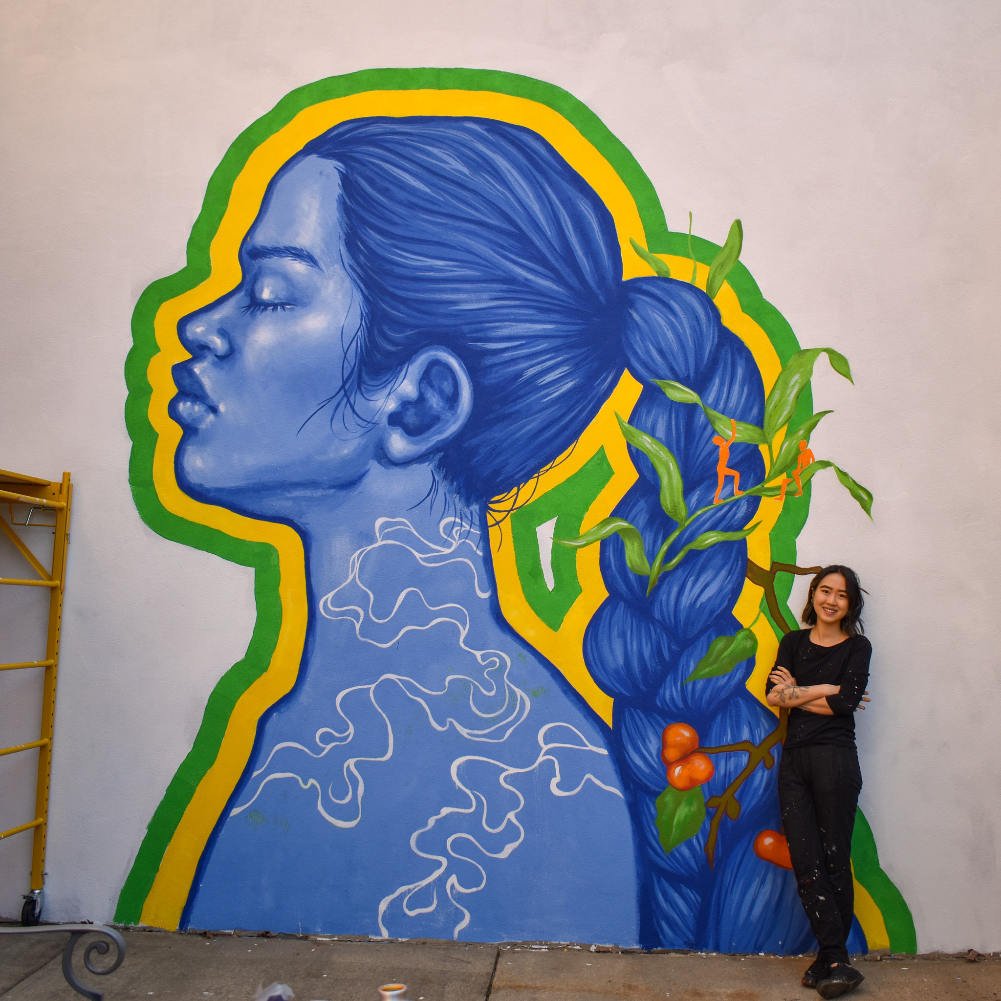 Artist Isabel Lu standing in front of part of the wall mural, depicting a woman standing in profile with a long braid behind her with seeds and grasses woven into her braid and small people climbing the grasses.
