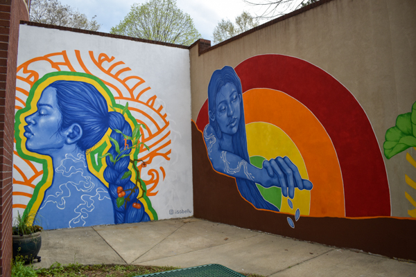 The mural spans two walls. On the back wall, a woman stands in profile with a long braid down her back and seeds and grasses woven into the braid. On the long wall to the right, another woman with long hair reaches out over the ground with her hand facing down, dropping seeds into the earth. Behind her is a rainbow.