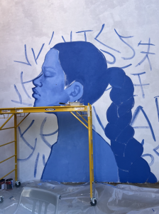 In the mural, a woman stands in profile, painted all in blue. She has a long braid behind her and around her portrait are shapes that look like letters or characters. Scaffolding sits in front of the mural.