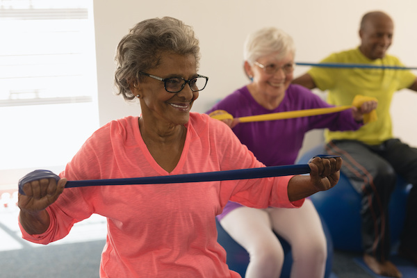 Older adults sit on exercise balls and stretch an exercise band between their arms while smiling