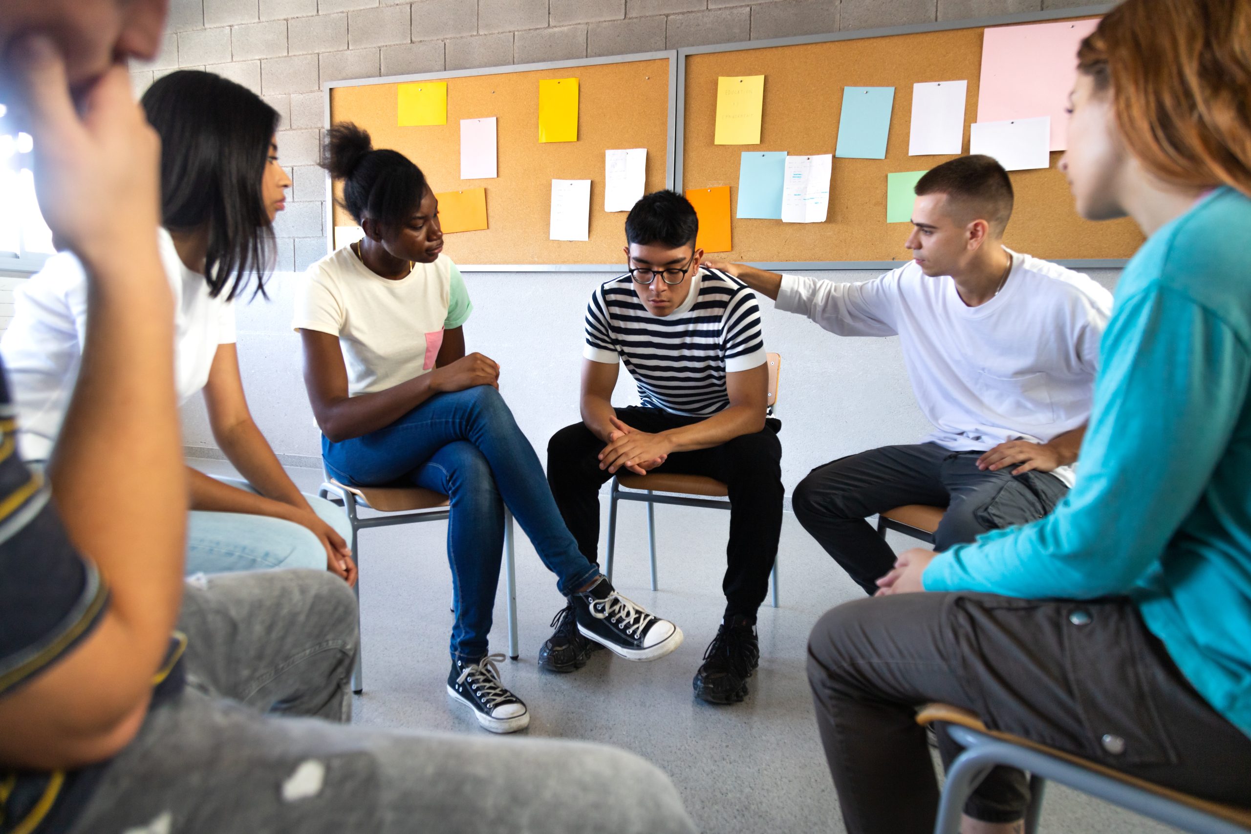 Sad hispanic male high school student sharing bad news with classmates. Multiracial teenagers sitting in circle in class comfort depressed teen boy. Mental health and education concepts.