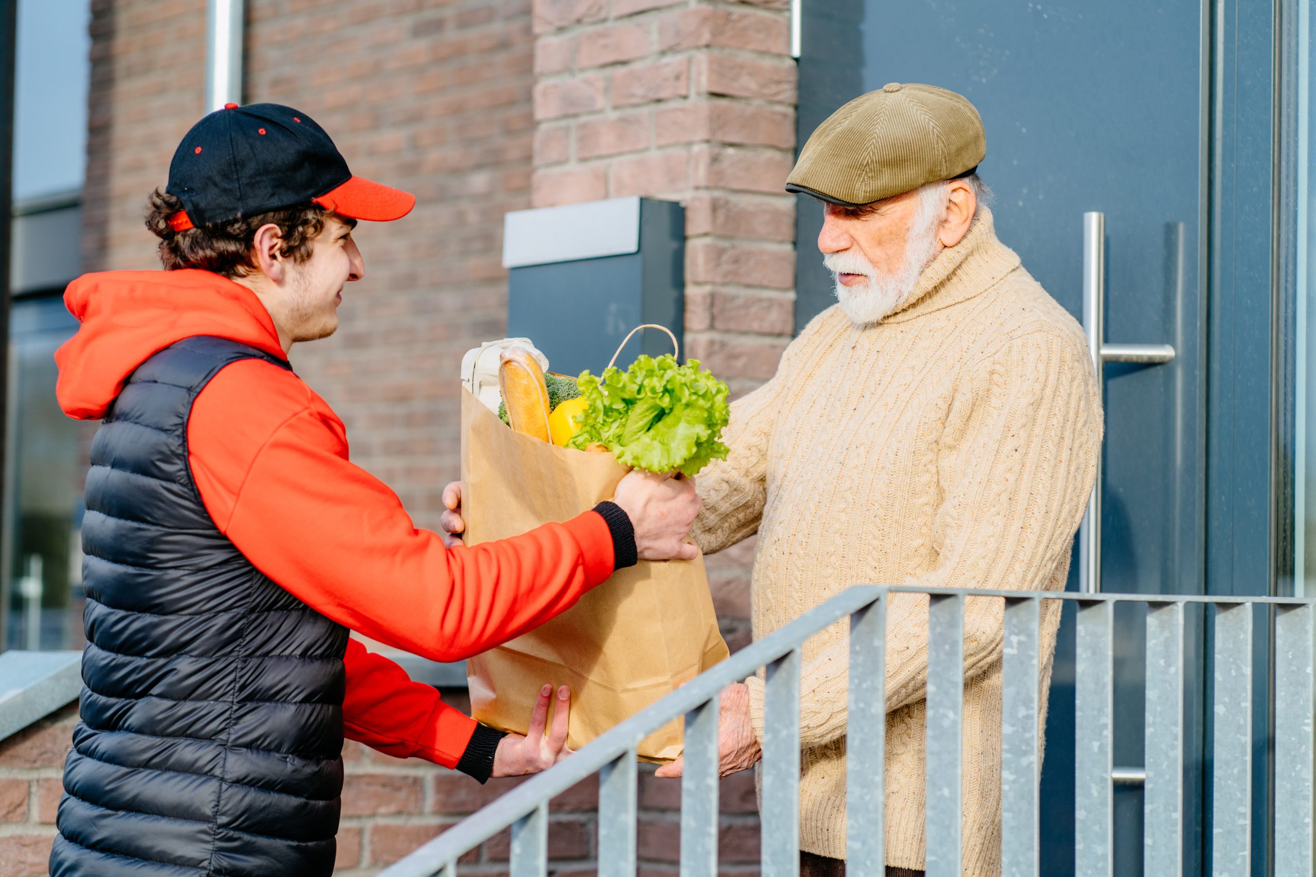 Profile shot of an elderly senior man taking to a food delivery male with a grocery box outdoor with building on background. Meal basket as social help and support.